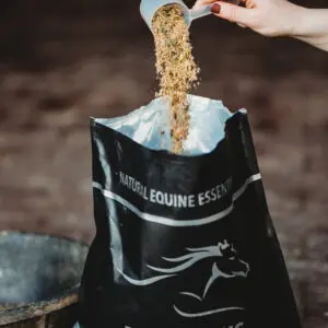A hand pours a scoop of multigrain Digest Plus supplement into an open Natural Equine Essentials bag, with the black packaging and logo clearly visible in a stable setting.