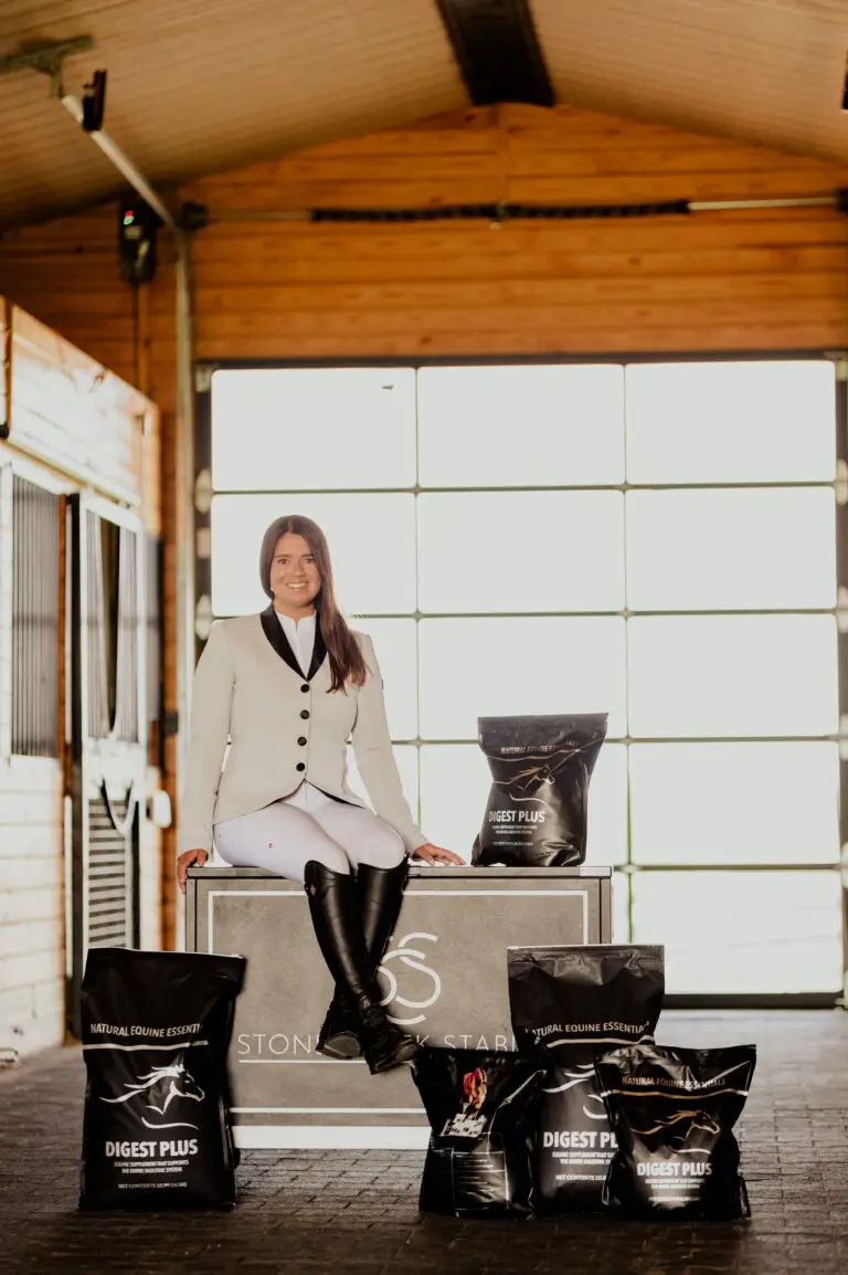 A woman in equestrian show attire sits on a Stone Creek Stables display box inside a stable, surrounded by several black bags of Digest Plus Natural Equine Essentials horse supplements.