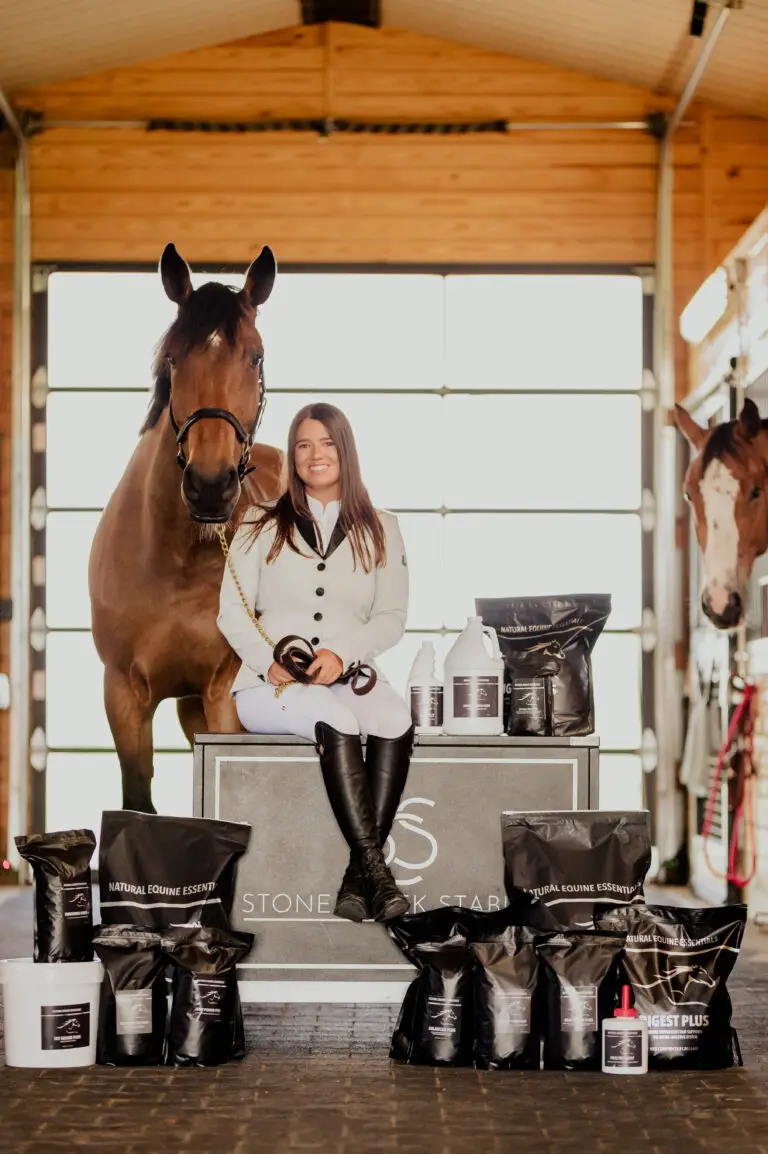 A smiling woman in a cream show jacket and tall boots sits on a Stone Creek Stables display box beside a bay horse, with a complete range of Natural Equine Essentials supplements and grooming products displayed around her.