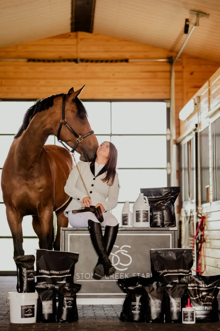 A smiling woman in equestrian attire sits on a Stone Creek Stables display box, looking up at a bay horse nuzzling her, surrounded by a full range of Natural Equine Essentials supplement and grooming products.