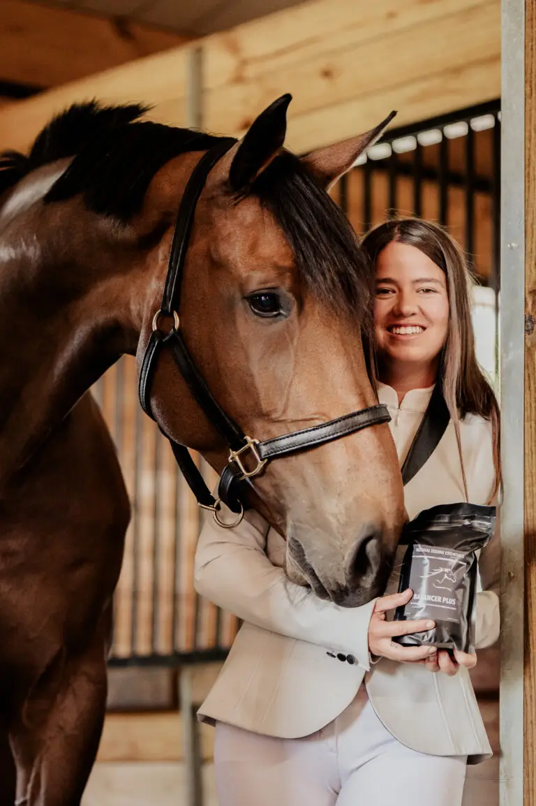 A smiling woman in a cream show jacket stands next to a bay horse in a stable, holding a black bag of Natural Equine Essentials Balancer Plus supplement.