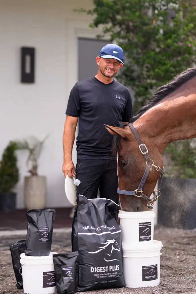 Man standing beside a brown horse eating from a white bucket, with Natural Equine Essentials supplement bags and buckets, including Digest Plus, displayed in front of a home setting.