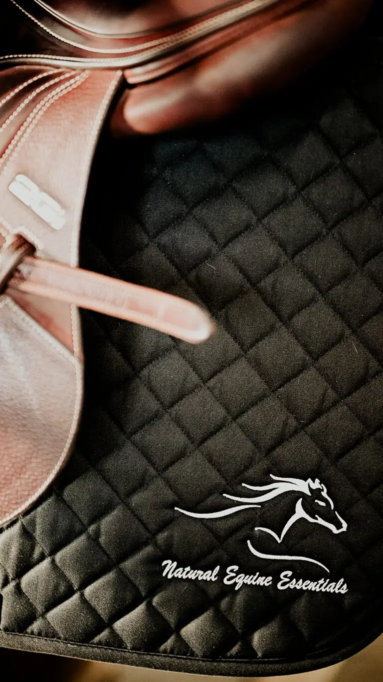 A close-up of a black quilted saddle pad embroidered with the Natural Equine Essentials logo, partially covered by a brown leather saddle.