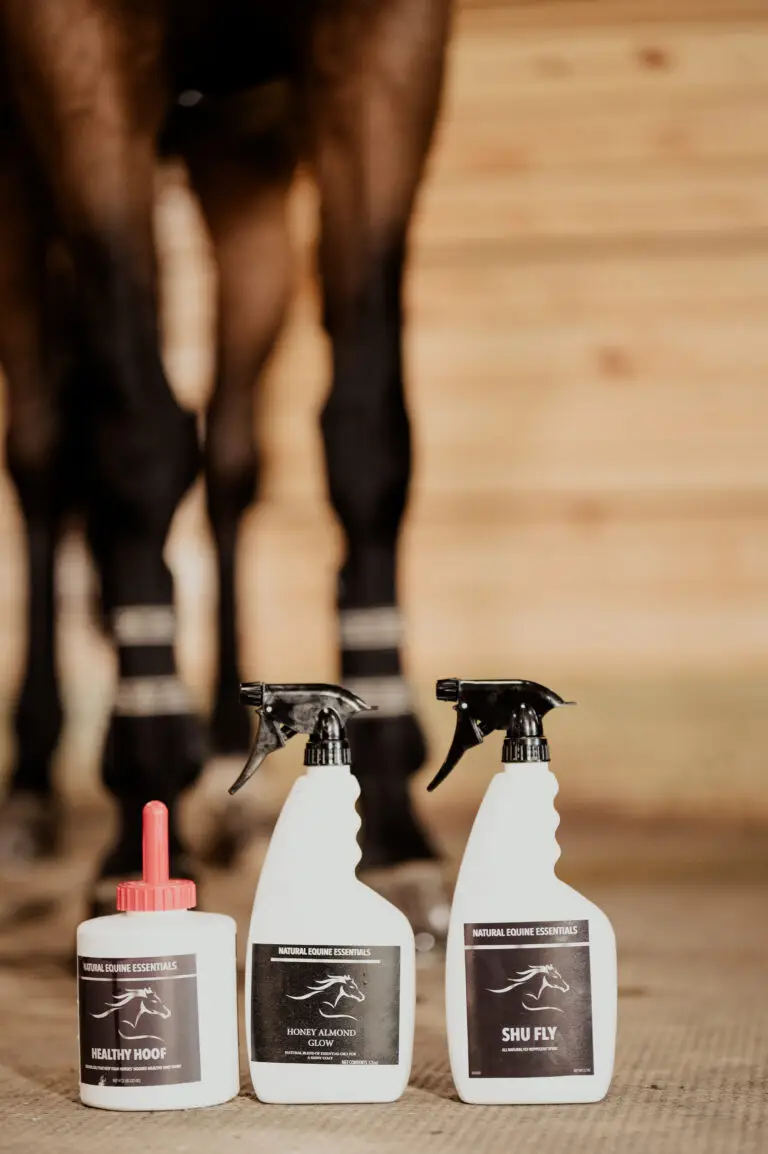 Three Natural Equine Essentials products, Healthy Hoof, Honey Almond Glow, and Shu Fly spray, are displayed on the ground in front of a horse's legs in a stable.