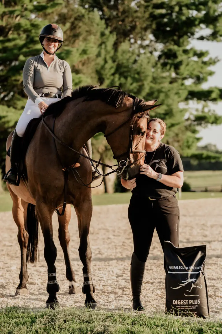 A woman in a riding helmet and breeches sits atop a bay horse while another woman stands beside them, affectionately holding the horse's face, with a black bag of Digest Plus Natural Equine Essentials supplements on the ground nearby.