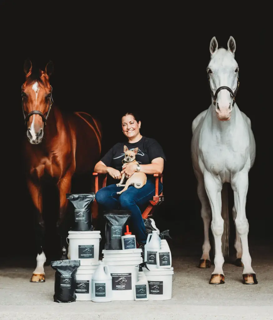 Woman sitting on a chair holding a small dog, positioned between a brown horse and a white horse, with multiple Natural Equine Essentials supplement buckets and pouches displayed in front against a dark background.