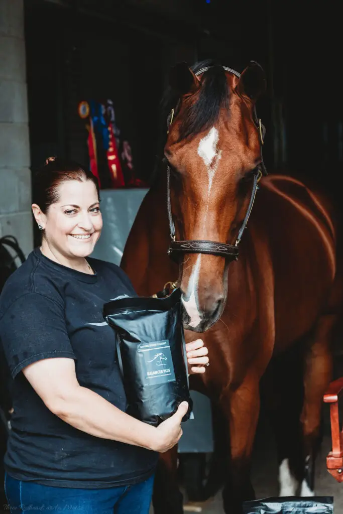 Woman standing beside a brown horse in a stable, holding a Natural Equine Essentials Balancer Plus supplement pouch while the horse leans toward it.