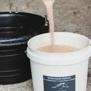 Hand pouring fine granular supplement from a white scoop into a white bucket labeled Natural Equine Essentials Electrolytes Plus beside a black feed tub in a barn aisle.