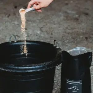 Hand pouring powdered Natural Equine Essentials Joint Power Plus supplement from a measuring scoop into a black feed tub, with the open product pouch standing beside it in a stable.