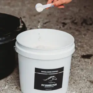 Hand holding a measuring scoop of powder above a white bucket labeled Natural Equine Essentials Fly Guard Plus, pouring supplement inside a stable setting.