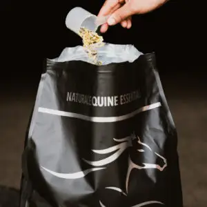 A hand pours a scoop of herbal equine supplement back into a Natural Equine Essentials Vita E Plus bag against a dark stable background.