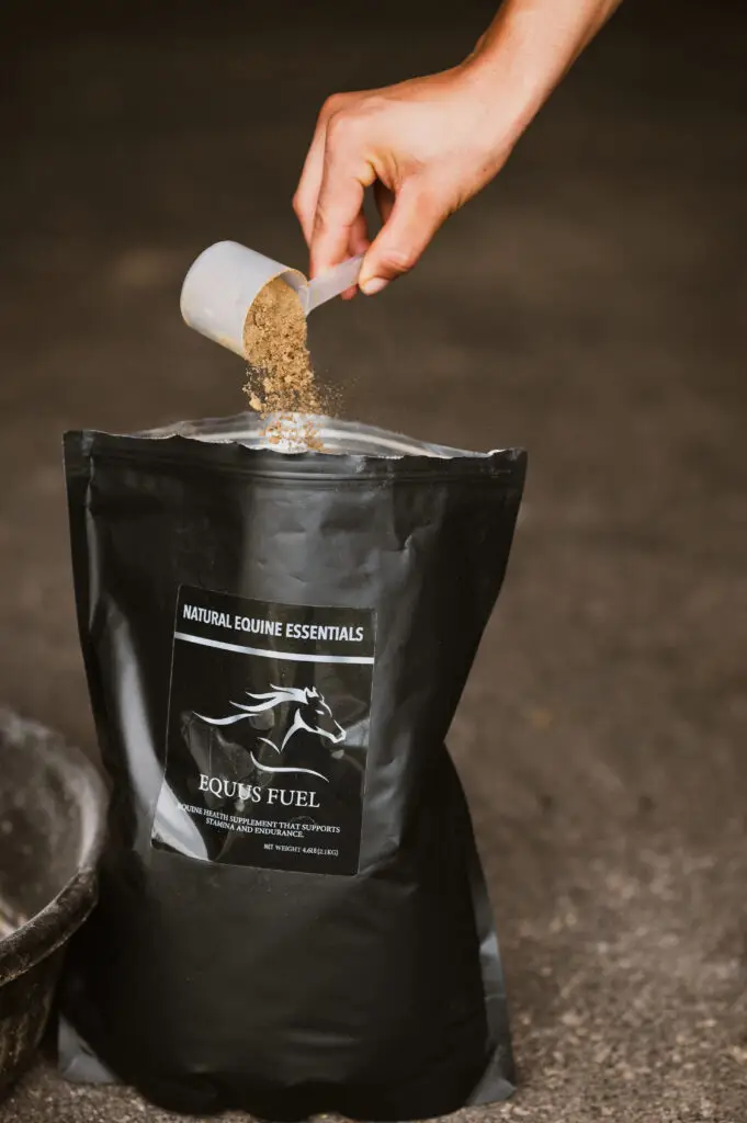 A hand pours a scoop of fine brown Equus Fuel supplement powder into an open Natural Equine Essentials bag, with the black packaging and logo clearly visible in a stable setting.