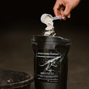 A hand pours a scoop of light-colored powder into an open black “Breath-EZ” Natural Equine Essentials supplement bag, with a feed bowl positioned nearby on the stable floor.
