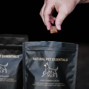 A hand holds a square dog supplement chew above a Natural Pet Essentials Joint Power Plus K-9 bag, with another supplement pouch partially visible beside it against a dark background.