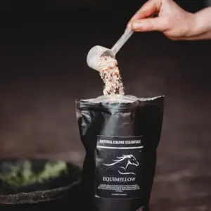 A hand pours a scoop of powder into an open black “Equimellow” Natural Equine Essentials bag, with fine particles falling into the pouch in a dim stable setting.