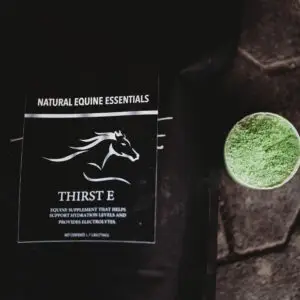 Close-up of a black Natural Equine Essentials “Thirst E” supplement bag lying on a stable floor beside a translucent scoop filled with bright green powder.