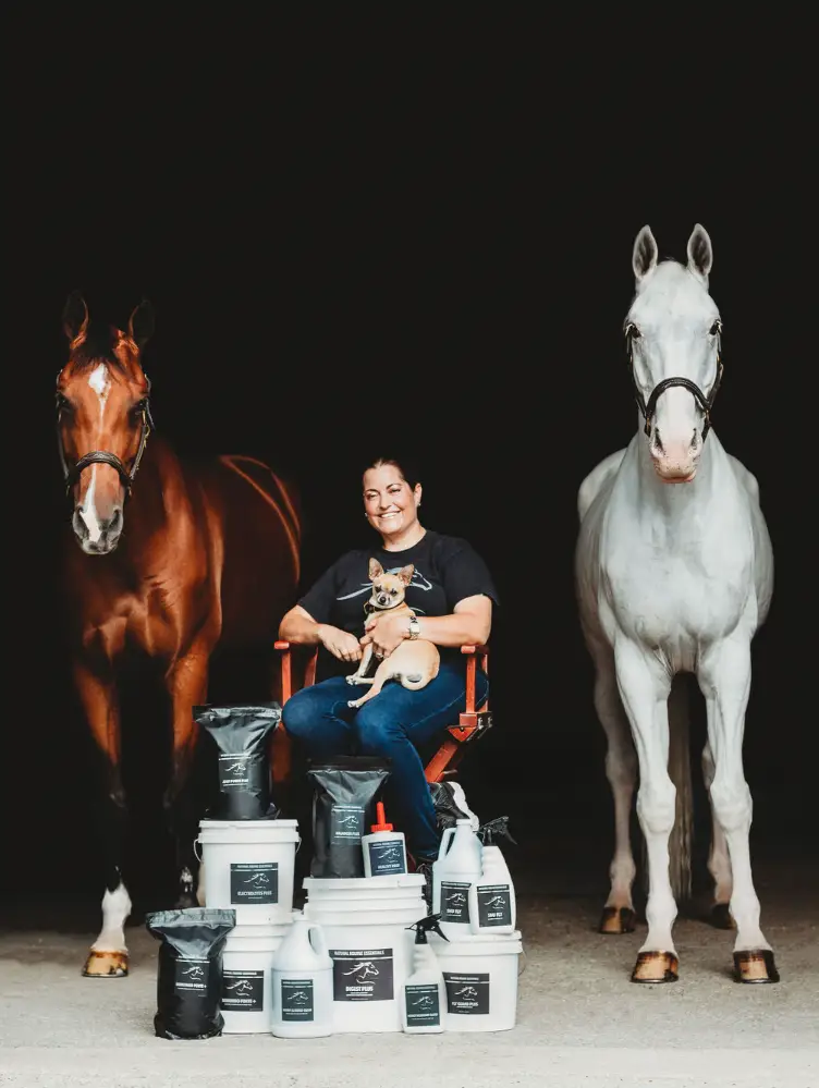 Woman sitting on a chair holding a small dog, positioned between a brown horse and a white horse, with multiple Natural Equine Essentials supplement buckets and pouches displayed in front against a dark background.