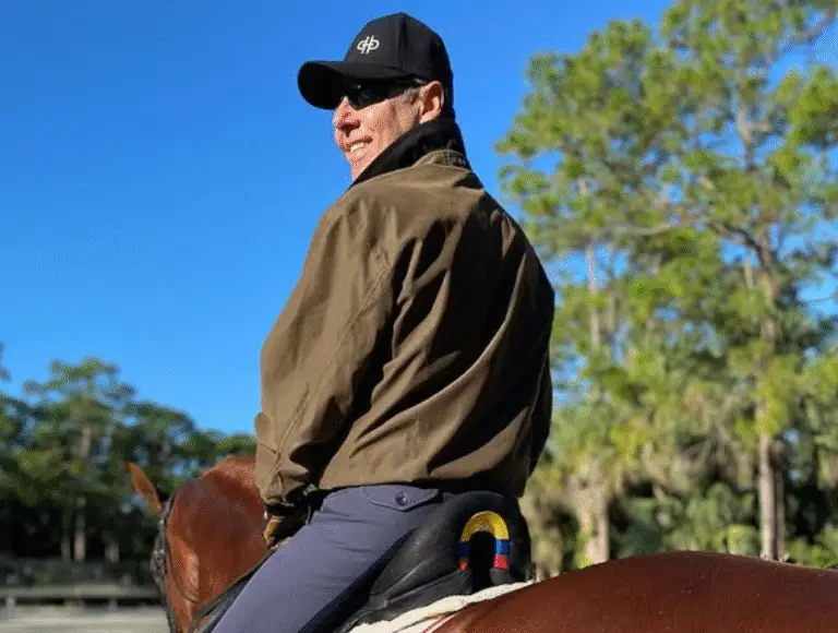A rider wearing a black cap, sunglasses, brown jacket, and tall riding boots sits on a chestnut horse in a sunlit sand arena, with trees and a clear blue sky in the background.