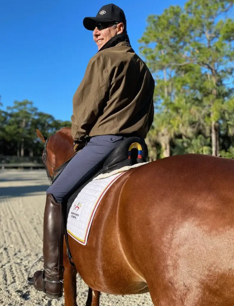 A rider wearing a black cap, sunglasses, brown jacket, and tall riding boots sits on a chestnut horse in a sunlit sand arena, with trees and a clear blue sky in the background.