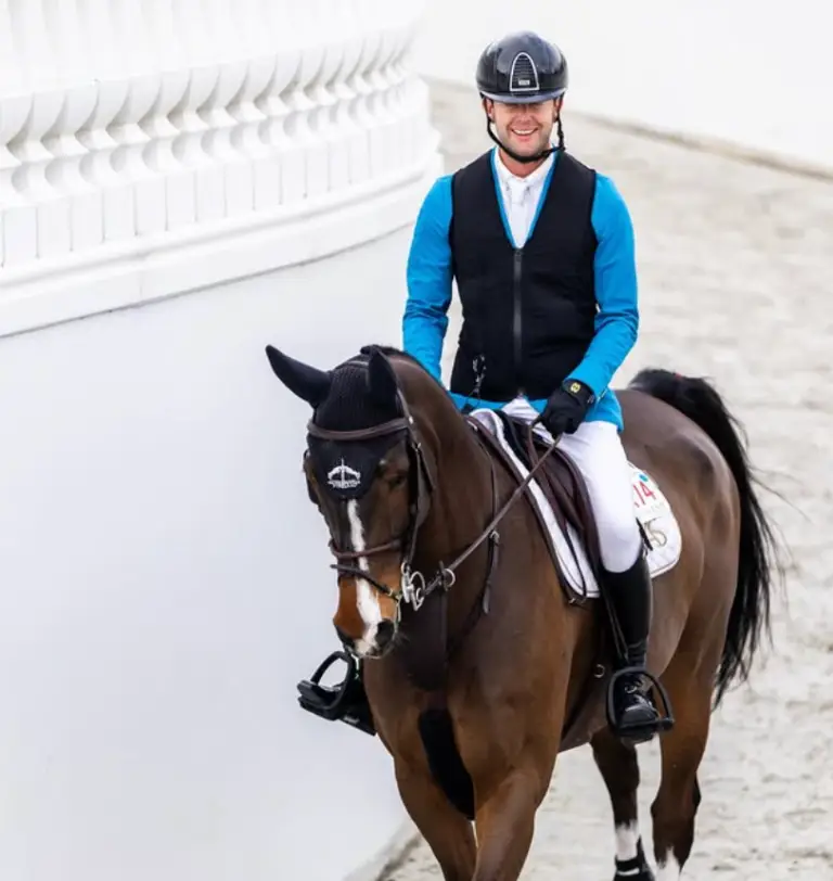 Professional show jumper Samuel Hutton riding a bay horse with a white blaze, wearing a blue competition jacket, black safety vest, and helmet at an equestrian event.