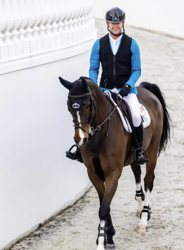 Professional show jumper Samuel Hutton riding a bay horse with a white blaze, wearing a blue competition jacket, black safety vest, and helmet at an equestrian event.